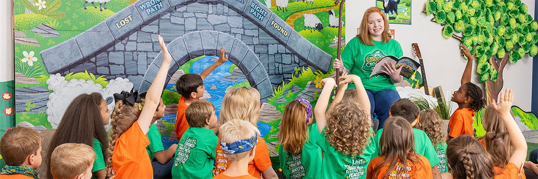 Children raising their hands as a teacher reads to them in front of a colorful VBS bridge mural